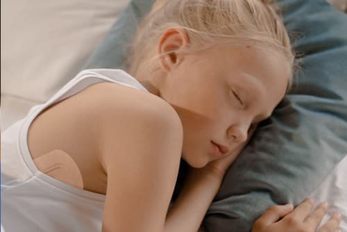 A young, blonde girl is asleep in bed, a STEADYTEMP® patch is visable under her arm.